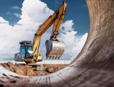 A powerful caterpillar excavator digs the ground against the blue sky. Earthworks with heavy equipment at the construction site. View from a large concrete pipe