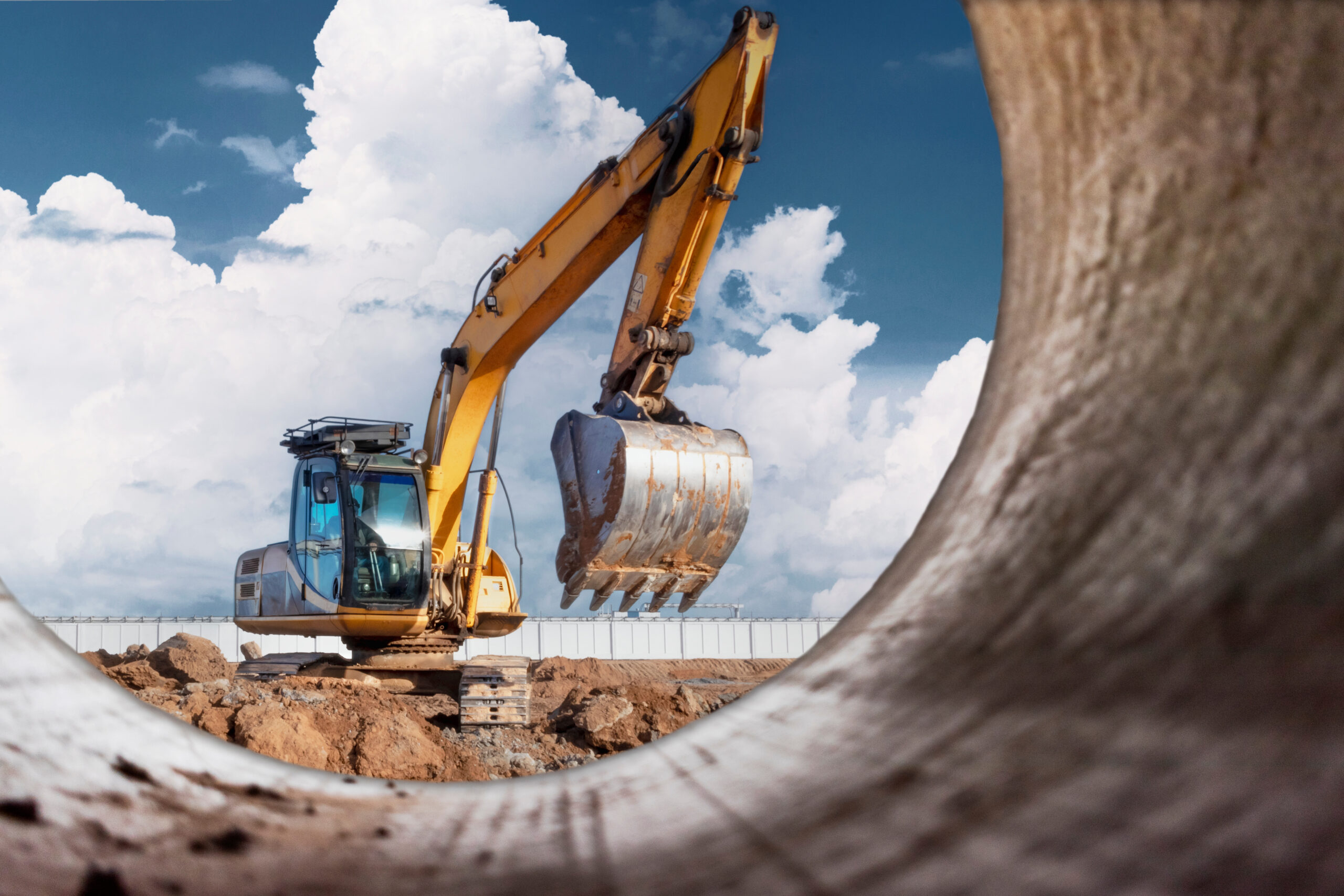 A powerful caterpillar excavator digs the ground against the blue sky. Earthworks with heavy equipment at the construction site. View from a large concrete pipe